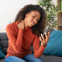 Eine Frau sitzt mit Nackenschmerzen auf dem Sofa und schaut auf ihr Handy