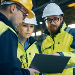 Drei Mitarbeitende in Sicherheitskleidung und Schutzhelmen diskutieren Fakten auf einem Tablet in der Maschinenhalle