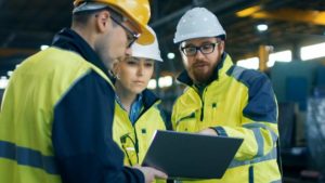 Drei Mitarbeitende in Sicherheitskleidung und Schutzhelmen diskutieren Fakten auf einem Tablet in der Maschinenhalle