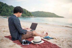 Junger Asiate sitzt auf einem Strandtuch am Strang mit dem Blick in Richtung Wasser, Laptop auf dem Schoß, neben sich Sonnenut und Digitalkamera