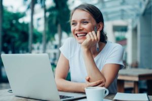 Junge digitale Nomading sitzt im Freien mit Laptop, Kaffeetasse und Stift bei der Arbeit und lacht, während sie den Blick schweifen lässt