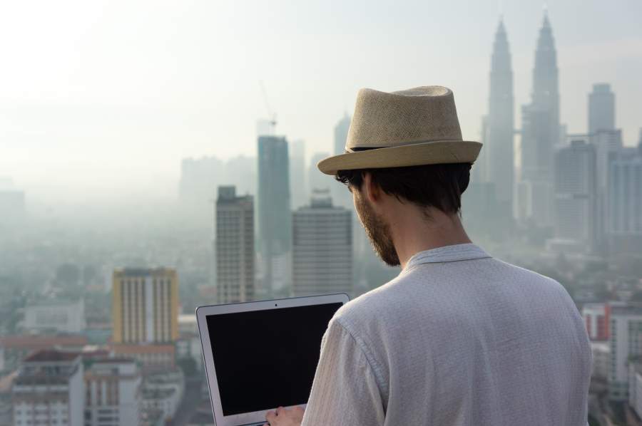 Junger Mann mit Sonnenhut arbeitet als digitaler Nomade auf dem Dach eines Hochhauses in urbaner Umgebung mit Hochhaus-Skyline im Hintergrund, er trägt einen Sonnenhut und weißes Shirt und arbeitet am Laptop, Rückenansicht