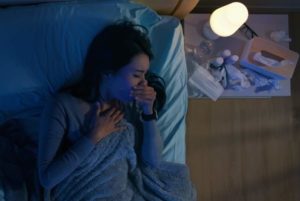 Eine Asiatische Frau liegt in einem halbdunklen Schlafzimmer seitlich unter mehreren blauen Decken in einem Bett und hält sich die Hand hustend vor das Gesicht während rechts neben ihr auf einem Nachtisch eine kleine weiße Nachtischlampe brennt, die neben verschiedenen Medikamentenpackungen und einer Taschentuchbox steht.
