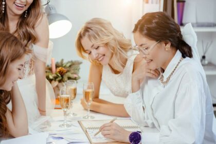 Drei junge, lachende Frauen sitzen mit einer Verkäuferin an einem Tisch auf dem vier Sektgläser stehen und suchen ein Brautkleid aus.