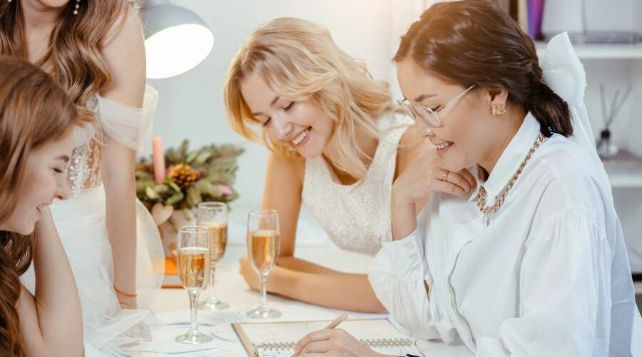 Drei junge, lachende Frauen sitzen mit einer Verkäuferin an einem Tisch auf dem vier Sektgläser stehen und suchen ein Brautkleid aus.