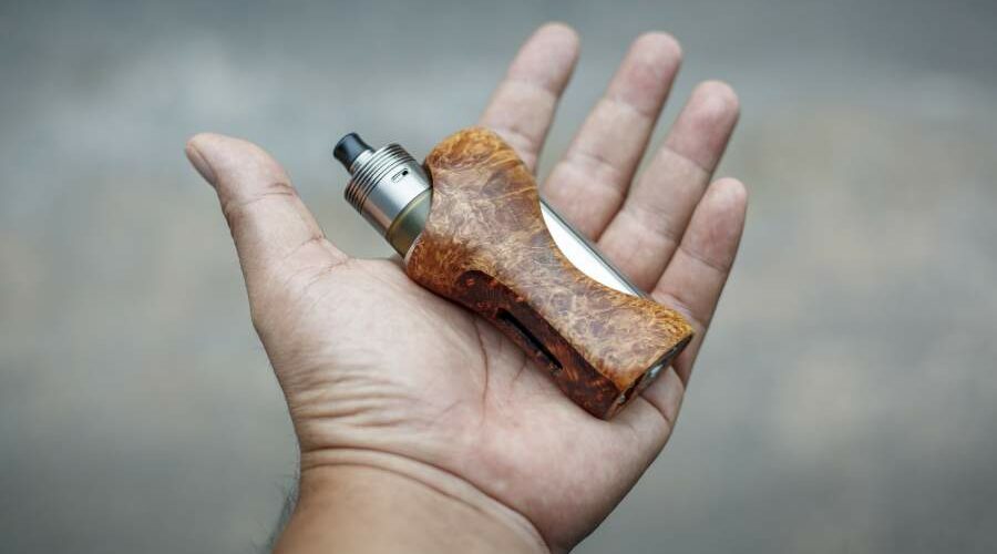 Ausgestreckte Hand vor grauem Hintergrund auf der ein moderner Vaporizer aus Holz liegt.