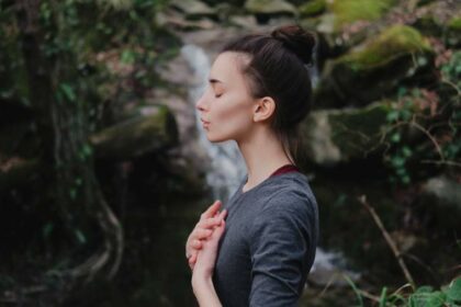 Junge Frau im Profil steht in der Natur und macht eine Atemübung, ein Wasserfall im Hintergrund