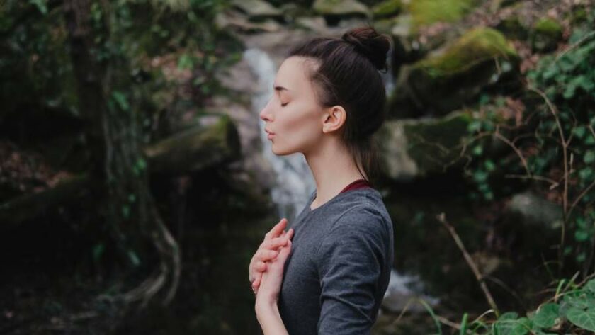 Junge Frau im Profil steht in der Natur und macht eine Atemübung, ein Wasserfall im Hintergrund