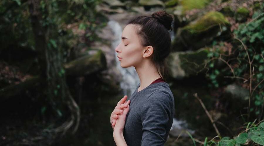 Junge Frau im Profil steht in der Natur und macht eine Atemübung, ein Wasserfall im Hintergrund