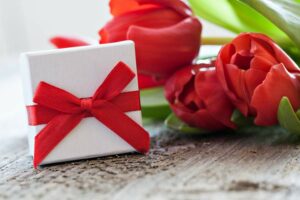 Kleine weiße Geschenkbox mit roter Schleife neben roten Rosen auf einem Holztisch