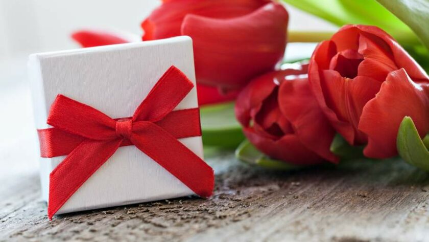 Kleine weiße Geschenkbox mit roter Schleife neben roten Rosen auf einem Holztisch