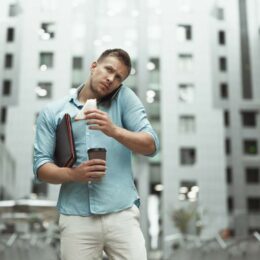 Junger, sportlich gebauter Mann in Hemd und heller Hose vor Bürogebäude wirkt gestresst, telefoniert unterwegs und hält ein angebissenes Sandwich, einen Coffee-to-go-Becher und eine Mappe in der Hand