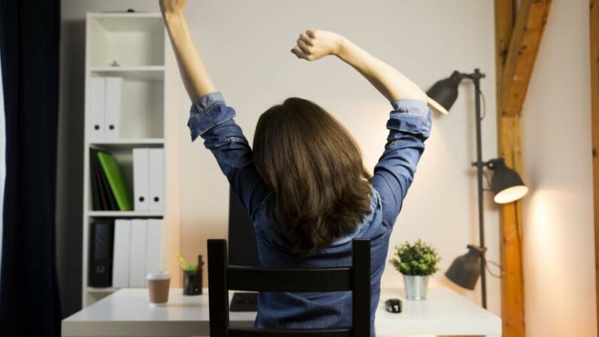 Rückensicht einer Frau, die sich streckt und im Homeoffice mit künstlichen Lichtquellen arbeitet