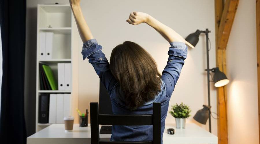 Rückensicht einer Frau, die sich streckt und im Homeoffice mit künstlichen Lichtquellen arbeitet