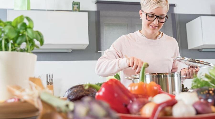 Junge Frau rührt in einem großen Kochtopf in einer hellen Küche um, im Vordergrund Chilischoten, Knoblauch, Paprika, Zwiebeln, ein Topf frischer Basilikum