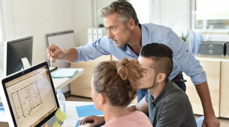 Ausbilder in Architekturbüro erklärt etwas am Grundriss auf dem Bildschirm einer Auszubilden und einem Auszubildenden