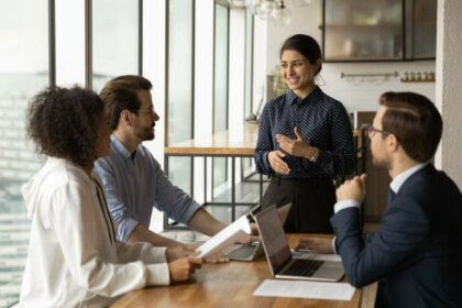 Vier diverse Personen in Business-Meeting, Frau mit indischen Wurzeln lächelt und gestikuliert