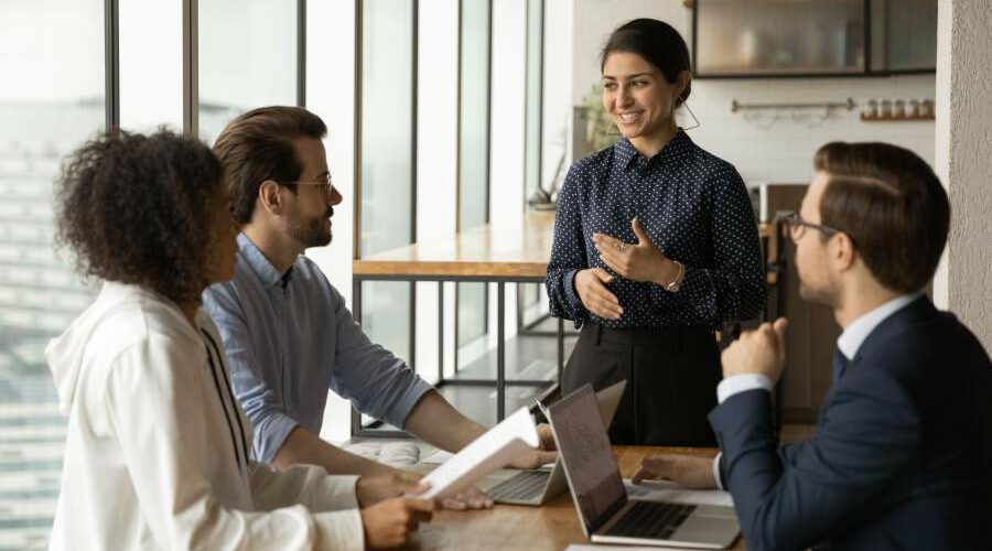 Vier diverse Personen in Business-Meeting, Frau mit indischen Wurzeln lächelt und gestikuliert