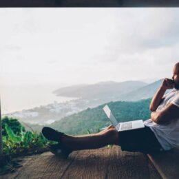 Junger Mann in Freizeitkleidung sitzt mit Laptop auf einer Veranda aus Holz und blickt nachdenklich in die Landschaft