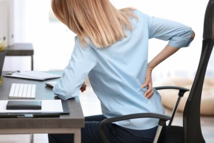 Junge Frau im Bürostuhl an Schreibtisch hält sich den Rücken in schmerzverzerrter Körperhaltung