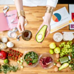 Vogelperspektive auf die Hände einer Ernährungsberaterin, die eine Glaskanne Öl und eine halbe Avocado über einem Tisch halten, darauf liegen Unterlagen, ein Maßband und verschiedene Lebensmittel