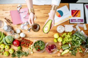 Vogelperspektive auf die Hände einer Ernährungsberaterin, die eine Glaskanne Öl und eine halbe Avocado über einem Tisch halten, darauf liegen Unterlagen, ein Maßband und verschiedene Lebensmittel