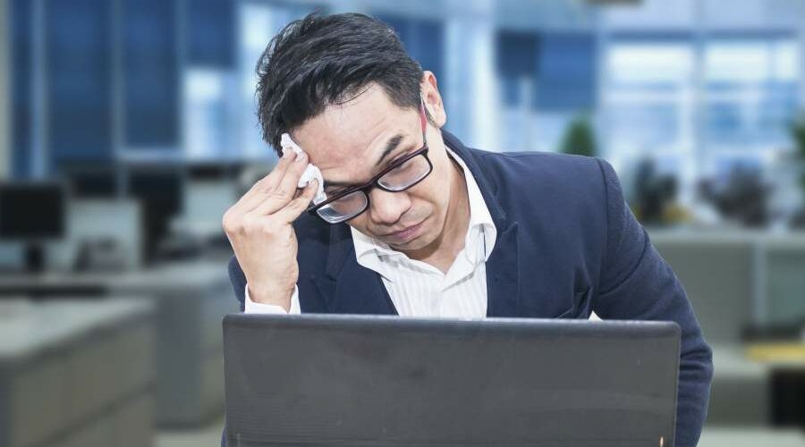 Geschäftsmann an Rechner in unbesetztem Großraumbüro blickt gestresst auf den Monitor und wischt sich den Schweiß von der Stirn