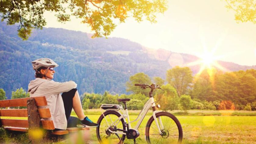 Frau mit Fahrradhelm und Sonnenbrille sitzt auf einer Bank in der Natur und in der Sonne, neben ihr steht ein Fahrrad