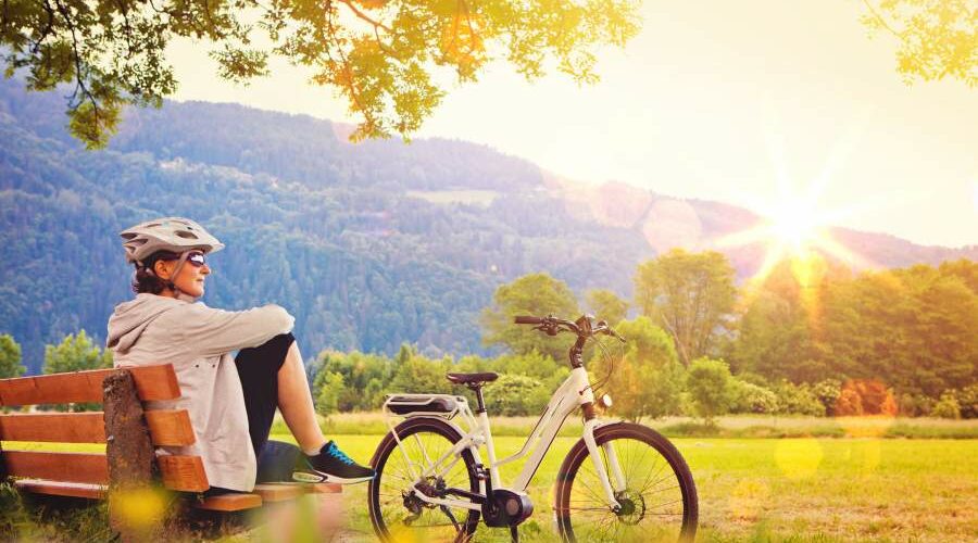 Frau mit Fahrradhelm und Sonnenbrille sitzt auf einer Bank in der Natur und in der Sonne, neben ihr steht ein Fahrrad