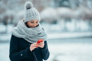 Junge Frau in Wintermütze, Schal und Mantel steht in verschneiter Landschaft und bedient ein Smartphone