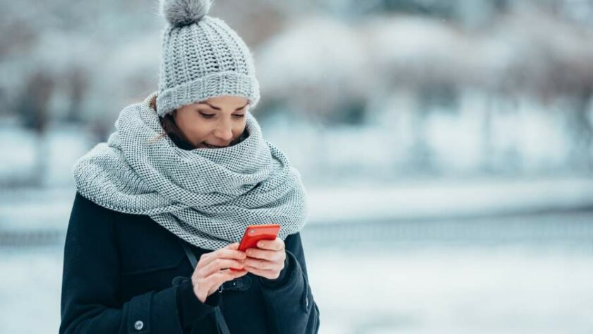 Junge Frau in Wintermütze, Schal und Mantel steht in verschneiter Landschaft und bedient ein Smartphone