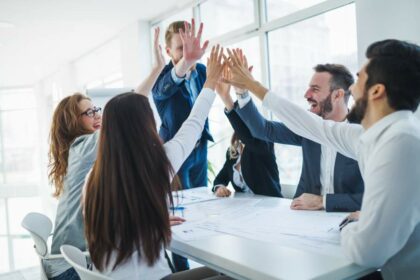 Motivierte Mitarbeitende an einem Meeting-Tisch schlagen die Hände in einem Gruppen-High-Five zusammen