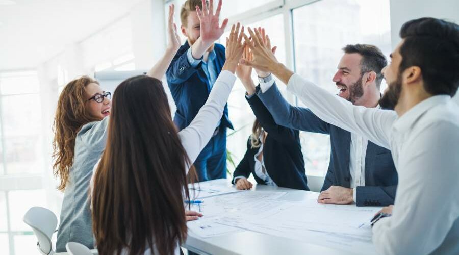 Motivierte Mitarbeitende an einem Meeting-Tisch schlagen die Hände in einem Gruppen-High-Five zusammen