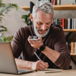 Mann Mitte 50 arbeitet im Homeoffice, telefoniert auf Lautsprecher und macht handschriftliche Notizen