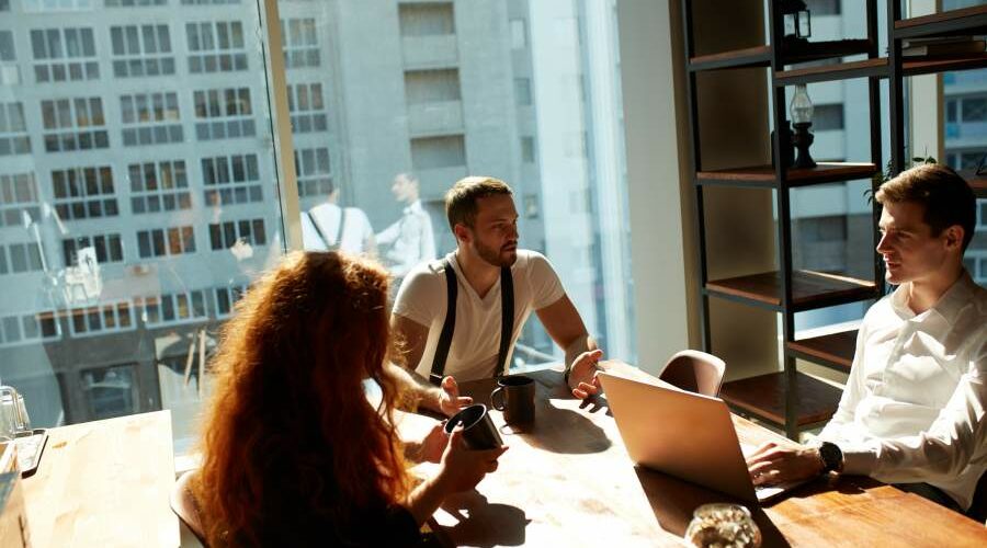 Junge Mitarbeitende im Team sitzen am Tisch während der Kaffeepause in einem hellen modernen Büroraum