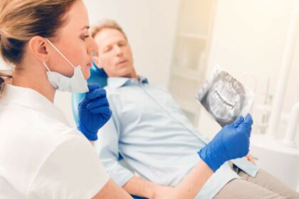 Zahnärztin betrachtet Röntgenaufnahme der Kiefer eines Patienten, der im Bildhintergrund auf dem Behandlungsstuhl in der Zahnarztpraxis sitzt