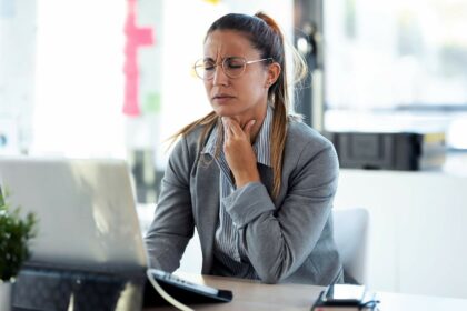 Junge Frau hat Erkältungssymptome und schlimme Halsschmerzen im Büro und fasst sich an den Hals